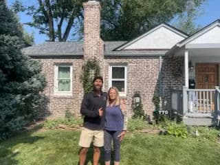 Couple standing in front of a brick house with green landscaping and trees.